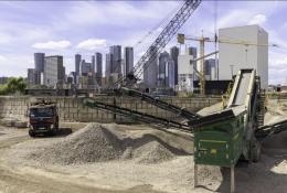 Piles of aggregate in a yard with a truck and green screen with a crane and tall buildings in the background