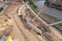 A large construction site by a river, including an orange crawler crane and other construction equipment