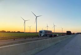 A truck on a motorway with a concrete safety barrier in the foreground and several wind turbines in the background.