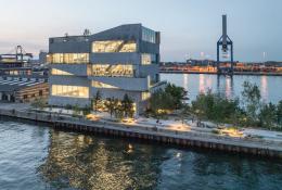 A seven storey concrete building next to a body of water with a ship loading crane in the background