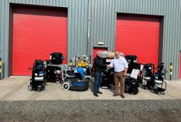 Two men shake hands infront of a range of floor grinding equipment, which is placed outside a grey warehouse with two, big red doors