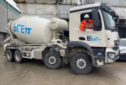 A white concrete mixer truck being driven by a man in red PPE