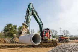 A green excavator with yellow pipe-handling grab moves a large piece of concrete pipe