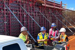 A group of construction workers in various colours of PPE stand in front of a wall of formwork