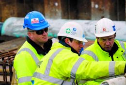 Three Costain construction workers in yellow PPE look at plans on a construction site.