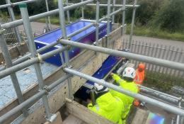 Two people in yellow PPE carry out a bridge inspection from a scaffold suspended from the side of the bridge
