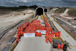 A large crowd of constructions workers in orange PPE stand by the entrance to the recently completed Chiltern Tunnel, HS2’s longest, which is now structurally complete following five years of construction. 