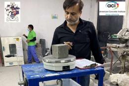 A man with a beard, waring a dark shirt inspects a concrete cube in a testing laboratory.