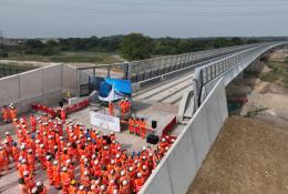 A large group of orange-clad PPE wearing construction workers gather at one end of the colne Valley Viaduct. Over 3,600 m³ of concrete was poured to form the kerb along the 3.4 km Colne Valley Viaduct – now structurally complete.
