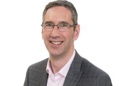 A head and shoulders shot of Peter Reekie of the Scottish Futures Trust will speak on sustainable infrastructure at the Glasgow Industry Day wearing glasses, an open necked pick shirt and a grey over-check jacket