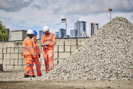 Three people in orange PPE stand discussing aggregate needs in a construction materials yard with a wall and city scape in the background