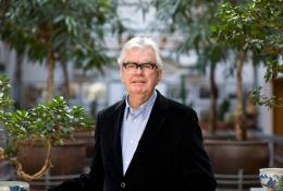 A grey-haired Sir Terry Farrell (1938-2025) wearing glasses, a dark jacket and light blue shirt photographed in front of an avenue of small trees in an urban setting © richard gleed / farrell