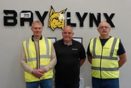 Three men stand in front of the BayLynx logo wearing PPE