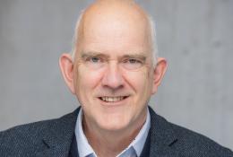 Head and shoulders photo of a bald, smiling man in open necked shirt, jumper and jacket