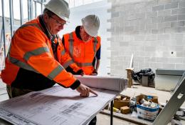 Two construction workers look at plans laid out on a table inside an under construction building