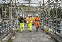 A group of construction workers walk along a concrete walkway, through a "tunnel" of scaffolding, wearing a variety of PPE , including orange hi vis jackets, yellow trousers, white hard hats and eye protectors