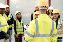 A group of youg people wearing yellow PPE and white and yellow hard hats listen to an older worker