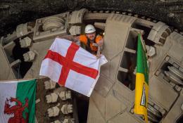 A man wearing a hard hat and waving a Cross of St George flag pokes his head through a TBM cutter wheel with a Welsh flag and another unidentified flag adorning the cutter head as HS2 engineers celebrate the breakthrough of tunnel boring machine Elizabeth at Washwood Heath in Birmingham, completing the 3.5-mile Bromford tunnel under the M6. (Image © HS2 Ltd)
