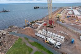 A red and yellow crawler crane lifts precast concrete elements at a shoreline construction site
