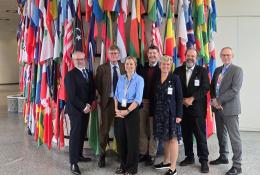 Four men and two women wearing work clothes stand in front of a large group of flags
