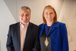 A smiling man and woman in business attire (dark suit, open necked shirt, blue jacket, black top) look at the camera