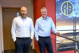 Two middle-aged men in smart, business casual wear stand on a stair landing in front of a large photo