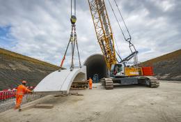 A yellow crawler crane lifts a precast concrete tunnel segment ready for placing on a cut and cover tunnel in the background