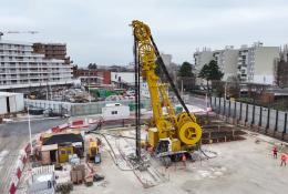 An elevated view of a yellow Soilmec SC-110 Urban cutter working on the Grand Paris Express, constructing 1,200mm-thick diaphragm walls up to 38 metres deep at Rueil–Suresnes Mont-Valérien station, surrounded by houses, low rise buildings and roads.