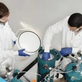 Two men in lab coats and safety glasses working in a lab next to two green canisters