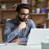A man with a beard and glasses sitting at a desk looking at a laptop