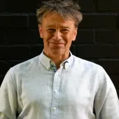 A head and shoulders picture of a man with unruly hair in a white shirt against a black background