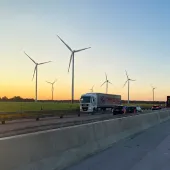 A truck on a motorway with a concrete safety barrier in the foreground and several wind turbines in the background.