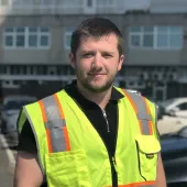 A man with short dark hair wearing yellow PPE in front of a mid-rise building