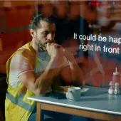 A man with dark hair in a top knot, wearing yellow PPE sitting at a cafe table looking anxious