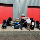 Two men shake hands infront of a range of floor grinding equipment, which is placed outside a grey warehouse with two, big red doors