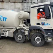 A white concrete mixer truck being driven by a man in red PPE