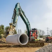 A green excavator with yellow pipe-handling grab moves a large piece of concrete pipe