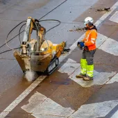 A man in orange, grey and yellow PPE uses a remote controlled piece of construction equipment fitted with a breaker to carry out repairs to a concrete road