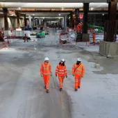 Three people in orange PPE walk across the competed base slab in the 850m long underground station box at Old Oak Common.