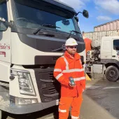 A man in orange PPE standing in front of a cement mixer holding a mobile device that uses the Podfather app to access their schedules and delivery notes, navigate routes, and record delivery details digitally. 