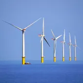 Six white and yellow wind turbines on a calm sea, part of the Walney Offshore Windfarm. Image: David Nixon / Creative Commons.