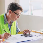 A man in glasses, blue shirt and yellow PPE vest sits working at a desk.