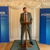 Mike Reader MP, the new chair of the All-Party Parliamentary Group for Excellence in the Built Environment, stands behind a podium, flanked by CIOB banners, speaking at the launch of the CIOB's Construction Client Guide: Leading Projects in the Built Environment, in Parliament last week.