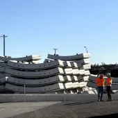 Two men in orange PPE stand next to a stack of precast concrete tunnel segments at the Western Harbour Tunnel Precasting facility in Emu Plains.