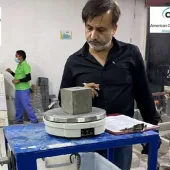 A man with a beard, waring a dark shirt inspects a concrete cube in a testing laboratory.