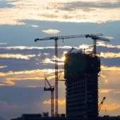 A high rise building framed against a sunset flanked by several tower cranes