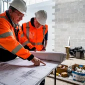 Two men in orange PPE look at plans on a table on a construction site