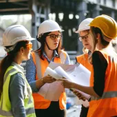 In the warm glow of the afternoon sun, a team of construction professionals gather closely, passionately engaged in a project discussion. Clad in safety helmets and high-visibility vests, they exchange ideas over blueprints, their collaboration a testament to the intricate planning that underpins successful infrastructure projects. Each individual's focused expression underscores the importance of each decision in the grand tapestry of urban development.