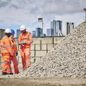 Three people in orange PPE stand discussing aggregate needs in a construction materials yard with a wall and city scape in the background