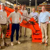 Five men dressed in smart shirts and trousers stand around a large red excavator arm